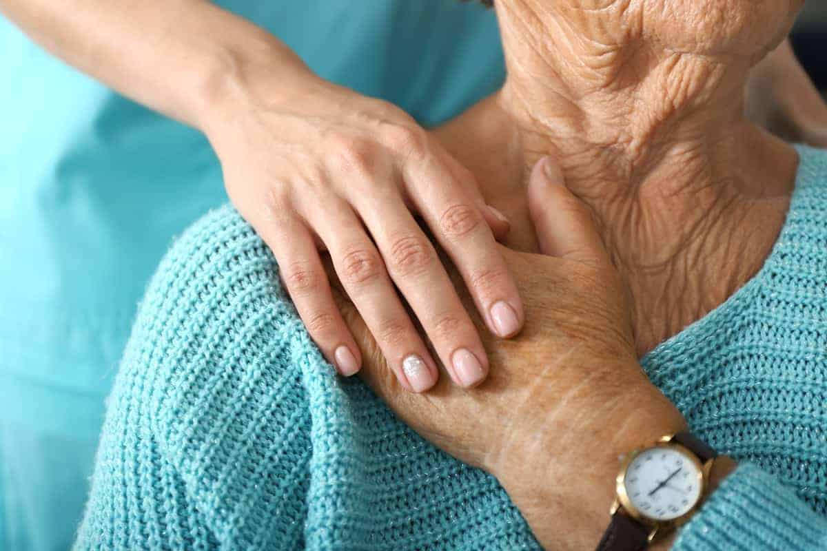 A caregiver resting her hand on an elderly woman's hand. The elderly woman is wearing a blue sweater