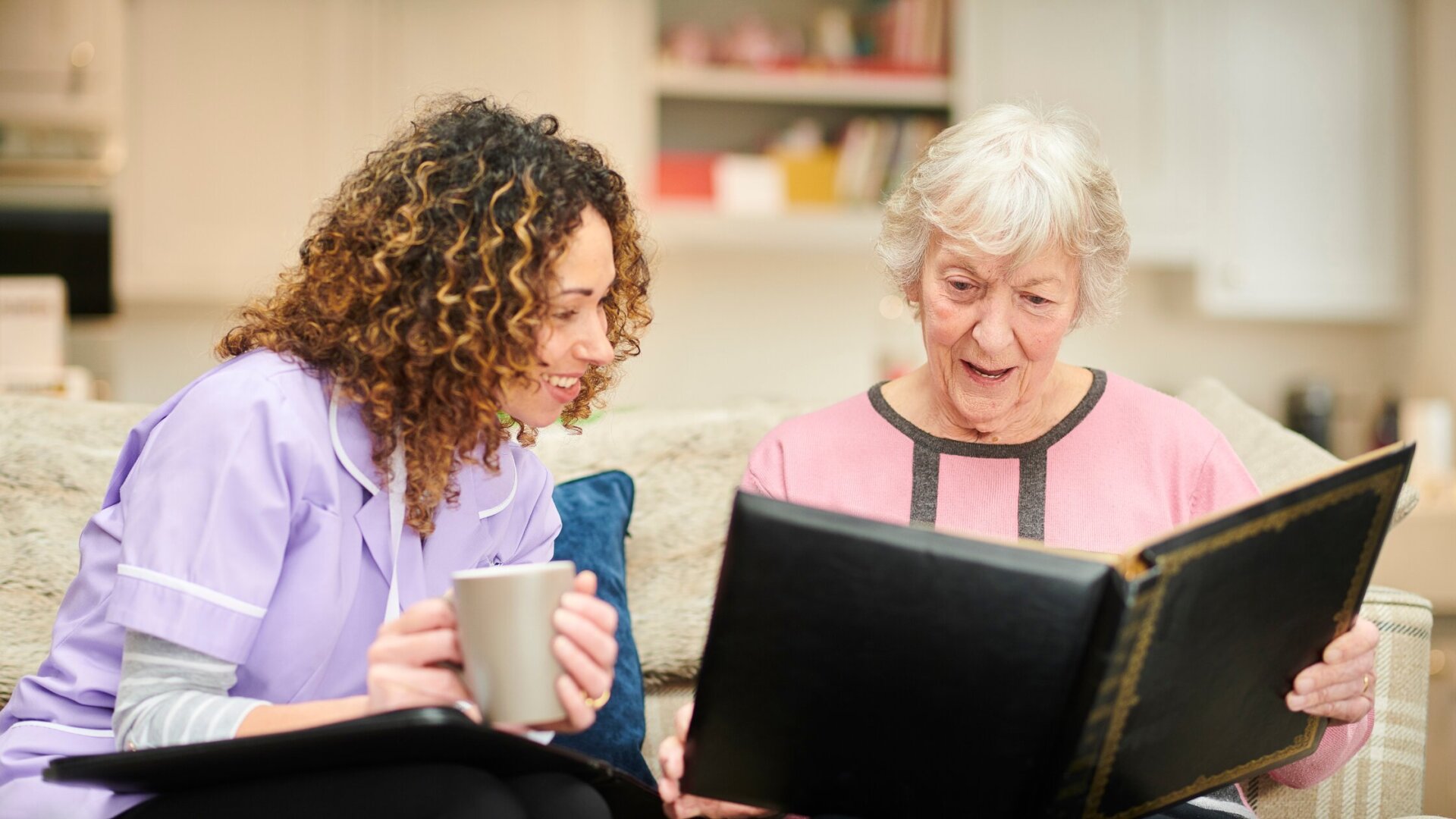 A caregiver and elderly woman sitting together on a couch looking at a photo book together