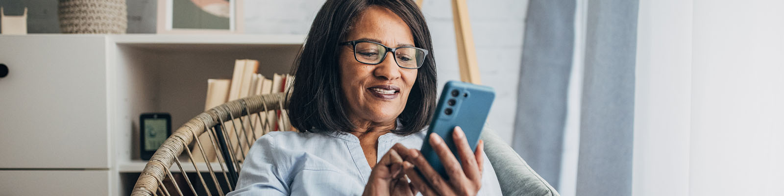 A senior woman sitting in a brown wicker chair looking down and scrolling on her smartphone